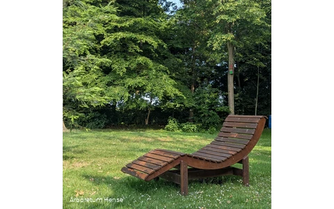 Eine Holzliege steht auf der Wiese im Vordergrund und im Hintergrund stehen einige Bäume