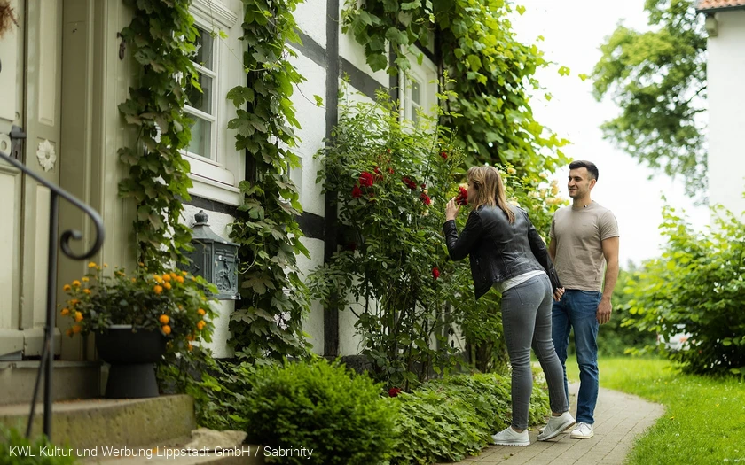 Eine Frau atmet den Duft einer Rose vor einem alten Fachwerkhaus am Stift Cappel im Lippstädter Ortsteil Cappel ein. Copyright: KWL Kultur und Werbung Lippstadt GmbH / Sabrinity