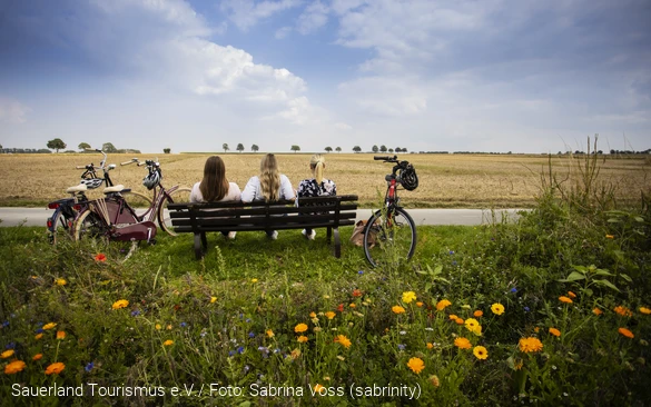 Drei Frauen sitzen auf einer Bank umgeben von Feldern. Sie blicken in die Ferne. Neben der Bank stehen Fahrräder. Foto: Sauerland Tourismus e.V./ Foto: Sabrina Voss (sabrinity)