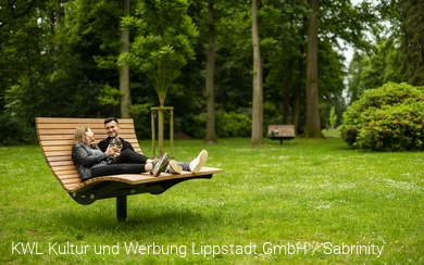 Ein Pärchen liegt auf einer großen Holzliege im grünen Kurpark. Sie trinken Wein. Foto: KWL Kultur und Werbung Lippstadt GmbH / Sabrinity