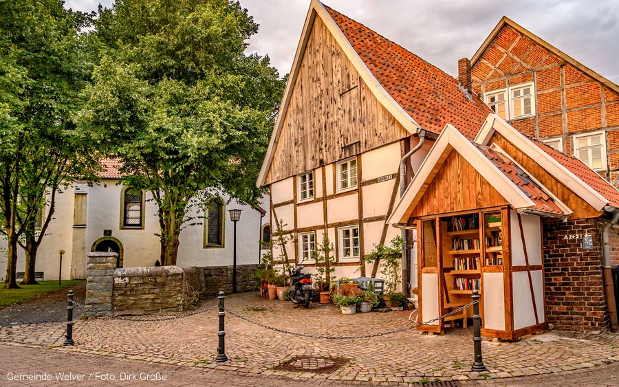 Alte, kleine Fachwerkhäuser in Kirchwelver. Im Hintergrund ist die Klosteranlage zu sehen. Im Vordergrund ist ein Bücher-Tausch-Schrank in Fachwerk-Optik. Copyright: Gemeinde Welver Foto: Dirk Große