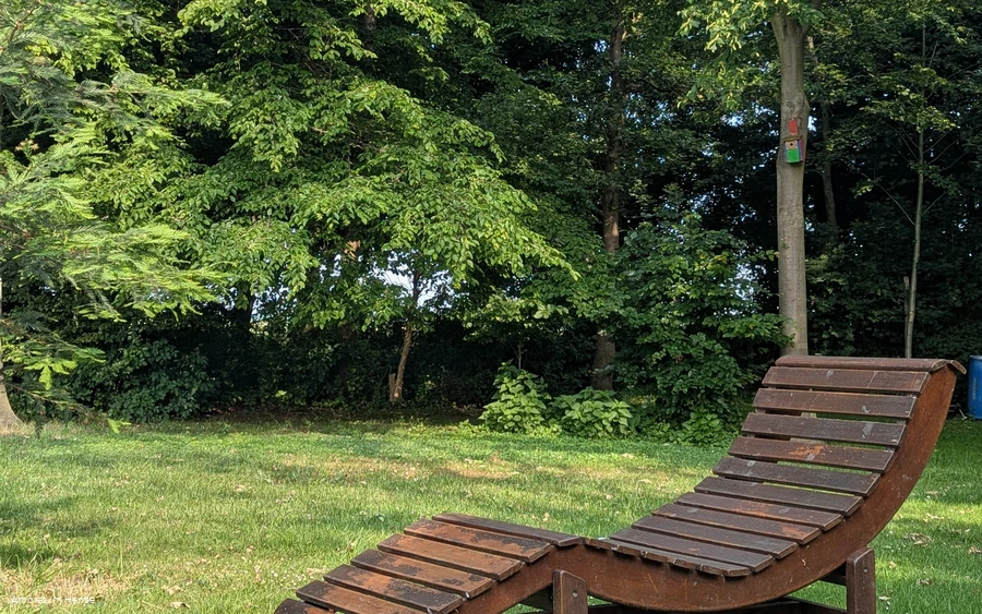 Eine Holzliege steht auf der Wiese im Vordergrund und im Hintergrund stehen einige Bäume