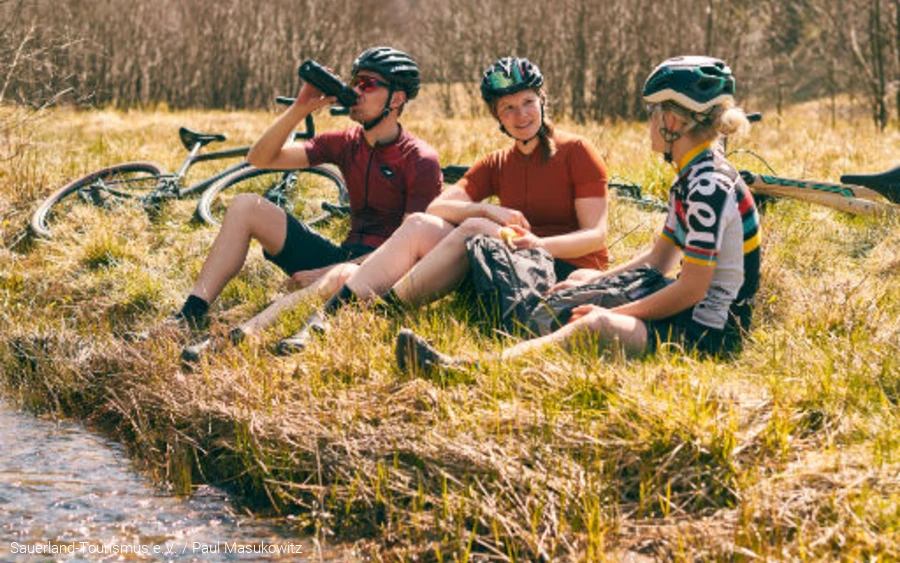 Drei Menschen machen Pause vom Radfahren. Sie sitzen an einem Bach in der Natur. Die Fahrräder liegen hinter ihnen. Foto: Sauerland-Tourismus e.V. / Paul Masukowitz