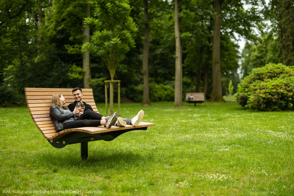Ein Pärchen liegt auf einer großen Holzliege im grünen Kurpark. Sie trinken Wein. Foto: KWL Kultur und Werbung Lippstadt GmbH / Sabrinity