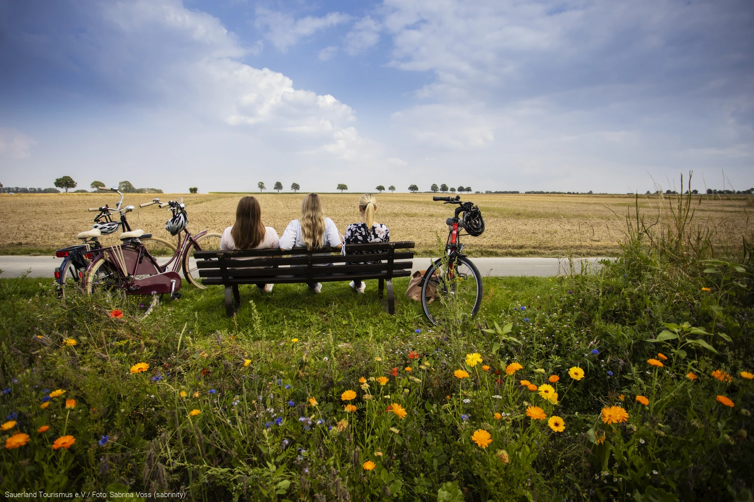 Drei Frauen sitzen auf einer Bank umgeben von Feldern. Sie blicken in die Ferne. Neben der Bank stehen Fahrräder. Foto: Sauerland Tourismus e.V./ Foto: Sabrina Voss (sabrinity)